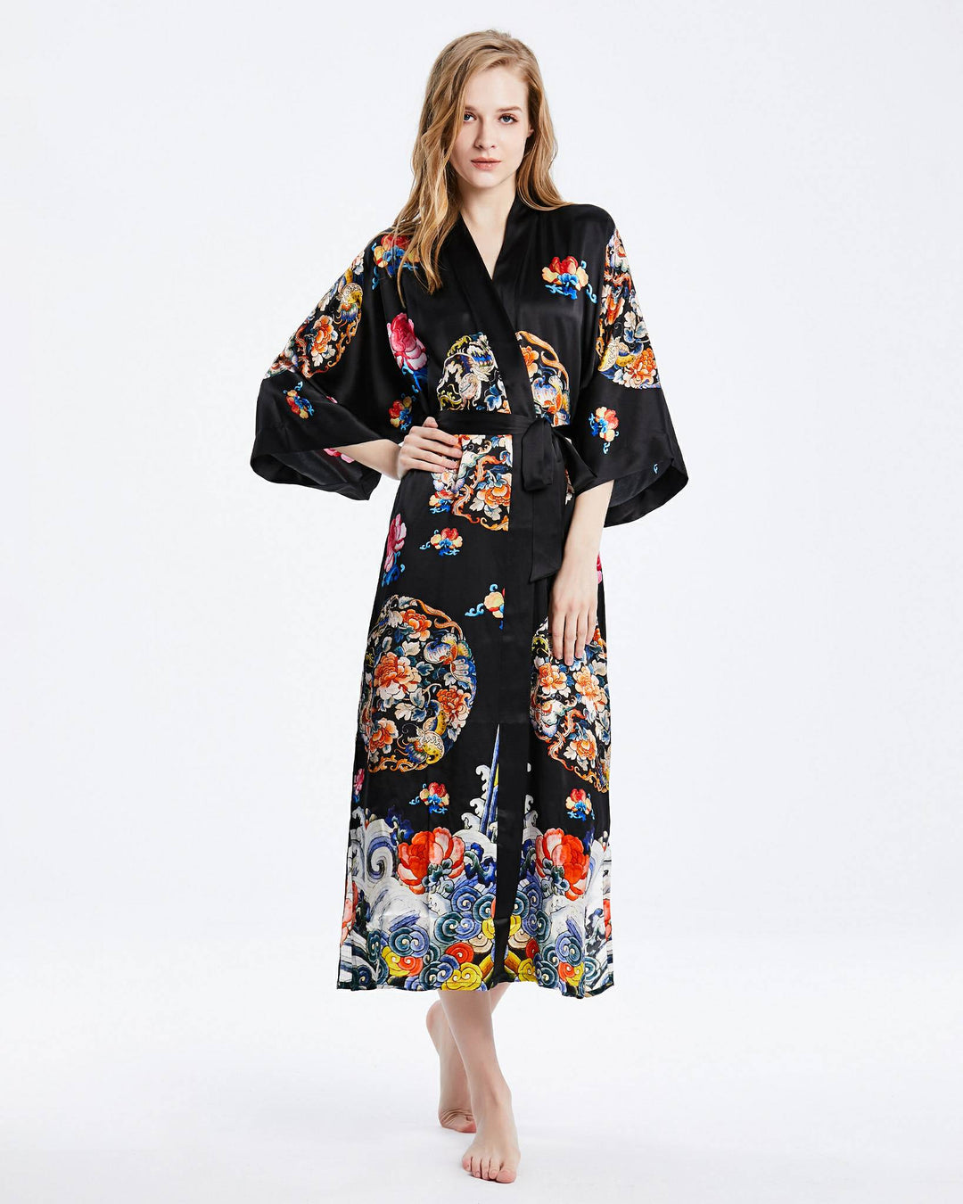 A woman stands barefoot wearing the Wheel of Fortune Long Silk Kimono by SusanSilk. This luxurious black floral kimono, adorned with colorful patterns on fine silk, complements her light hair and neutral expression.#color_black