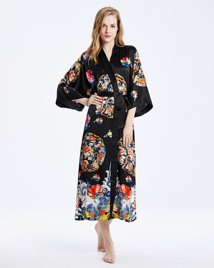 A woman stands barefoot wearing the Wheel of Fortune Long Silk Kimono by SusanSilk. This luxurious black floral kimono, adorned with colorful patterns on fine silk, complements her light hair and neutral expression.#color_black