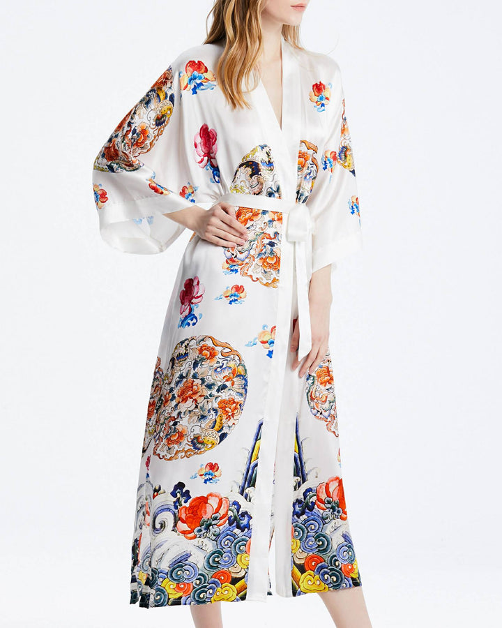 A woman wearing the Wheel of Fortune Long Silk Kimono by SusanSilk, adorned with colorful and intricate designs, stands with one arm slightly raised. The kimono features a tied belt at the waist.#color_white