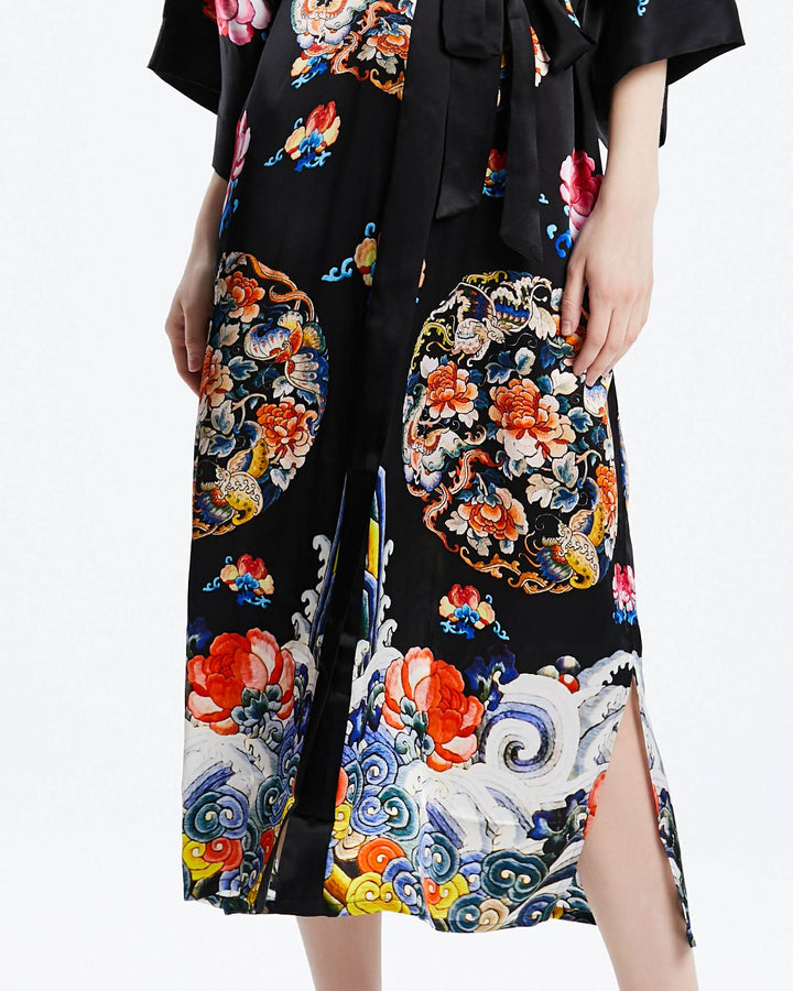 A person wearing the Wheel of Fortune Long Silk Kimono by SusanSilk, adorned with colorful floral and cloud patterns and featuring slits on both sides, is shown from the neck down.#color_black
