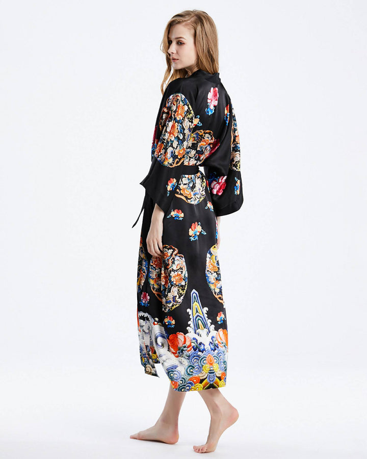 A woman stands barefoot, wearing the Wheel of Fortune Long Silk Kimono by SusanSilk, adorned with colorful floral and decorative patterns, looking over her shoulder.#color_black