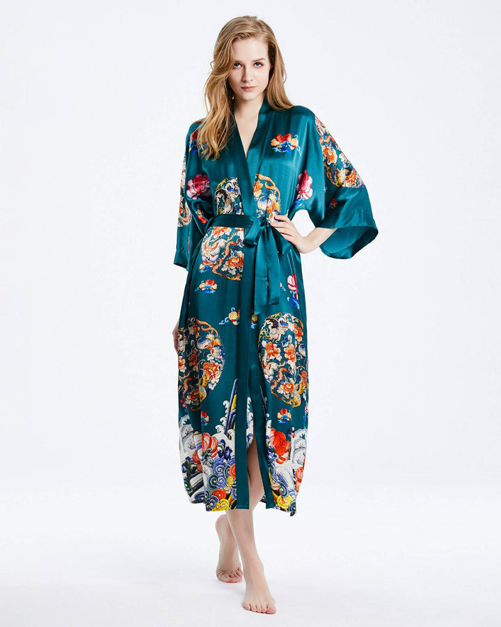 A person stands barefoot wearing a SusanSilk Wheel of Fortune Long Silk Kimono, featuring a teal, floral and colorful design. The kimono has a belt tied at the waist and long sleeves. The background is plain white.#color_sea-green