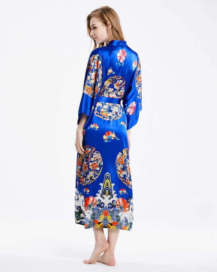 A woman stands barefoot, wearing the Wheel of Fortune Long Silk Kimono by SusanSilk, adorned with vibrant floral and geometric patterns, while facing partially away from the camera.#color_royal-blue