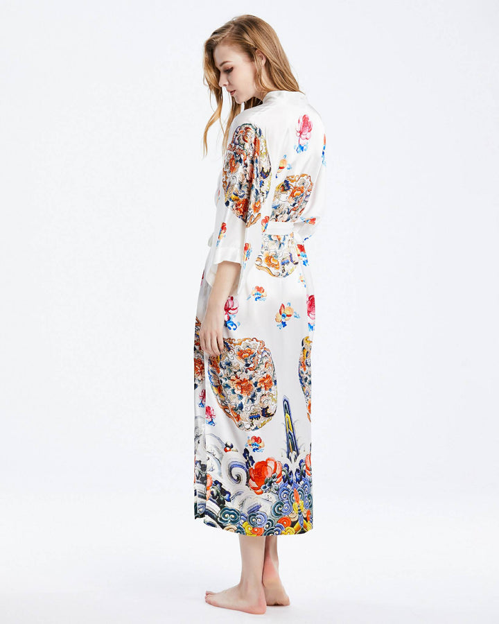 A woman stands barefoot in a Wheel of Fortune Long Silk Kimono by SusanSilk, featuring colorful floral and abstract patterns, facing slightly away from the camera.#color_white
