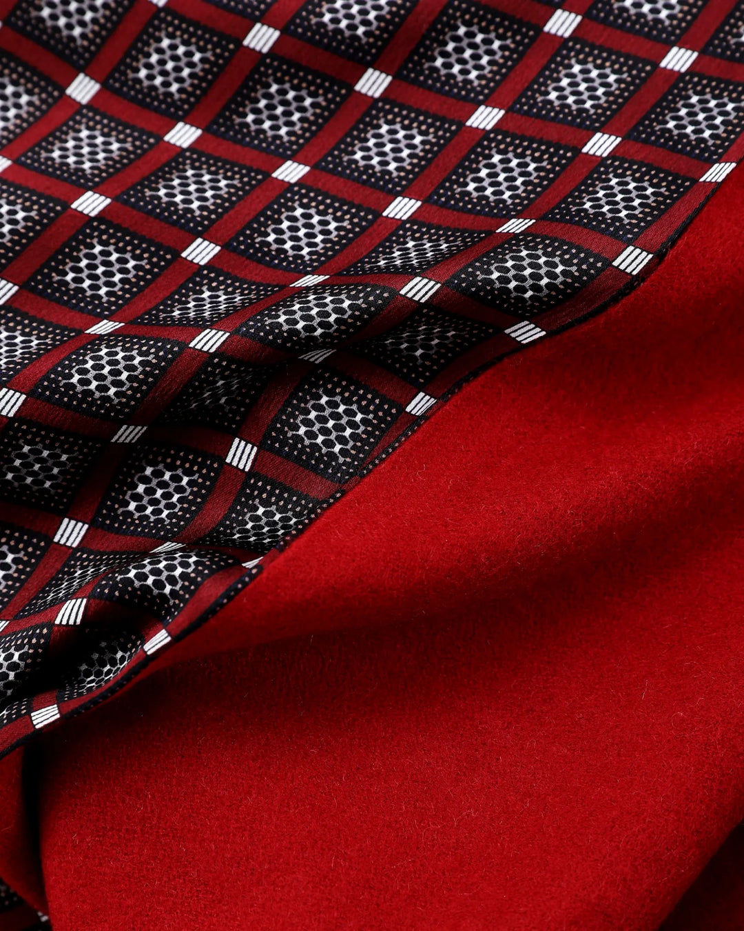 A close-up of a folded scarf showing two sides: one side features a red, black, and white geometric checkered pattern, and the other side is a solid deep red.#color_wine-red