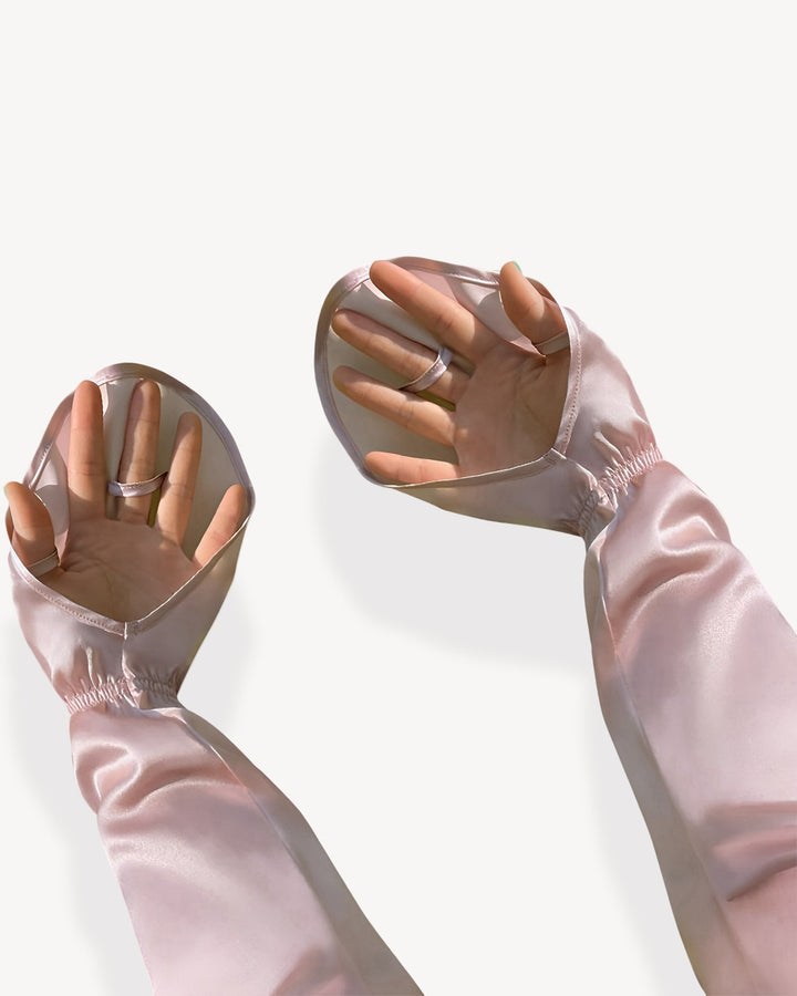 Two hands extended upward through sheer, light pink Sun Protection Silk Over Sleeves from SusanSilk, featuring thumb holes, set against a plain white background.