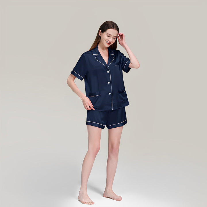 A person wearing the SusanSilk 19 Momme Short Silk Pajamas Set in navy blue, featuring white piping and short sleeves, standing barefoot against a plain background.