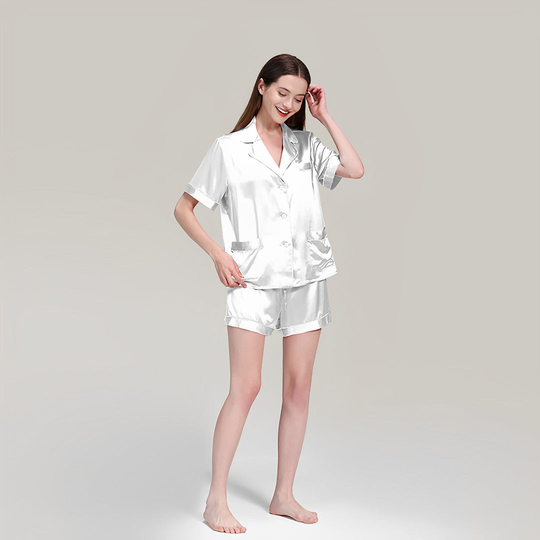 A woman wearing SusanSilk's 19 Momme Short Silk Pajamas Set in silver satin stands barefoot against a plain background, smiling and touching her hair.