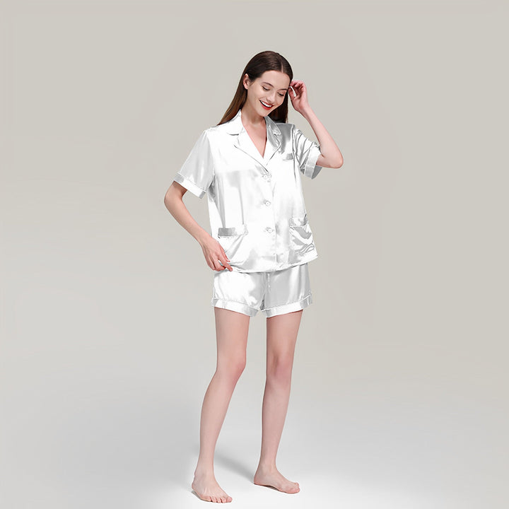 A woman wearing SusanSilk's 19 Momme Short Silk Pajamas Set in silver satin stands barefoot against a plain background, smiling and touching her hair.