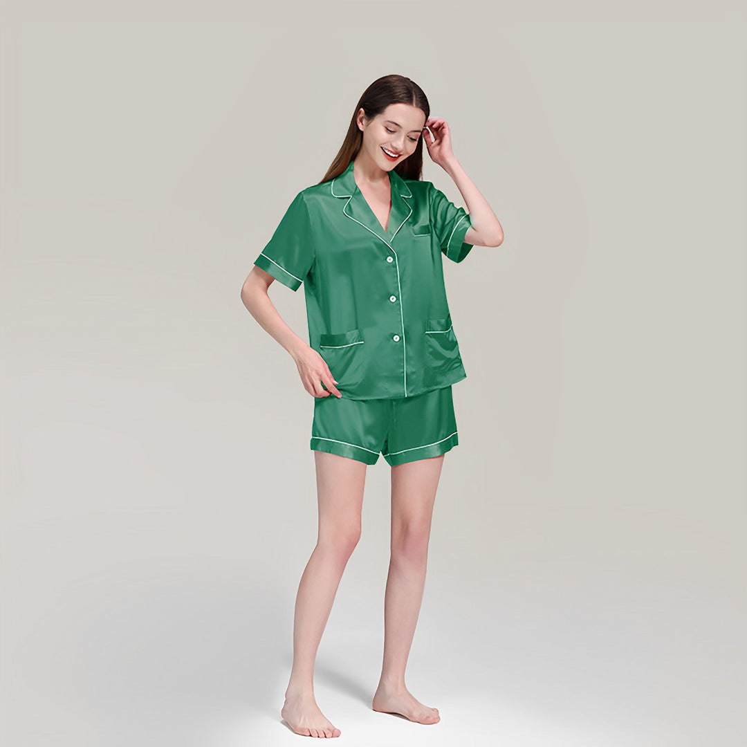 A person stands barefoot, wearing the SusanSilk 19 Momme Short Silk Pajamas Set in green, consisting of a button-down shirt and shorts. The person has long hair and is smiling while touching their head.