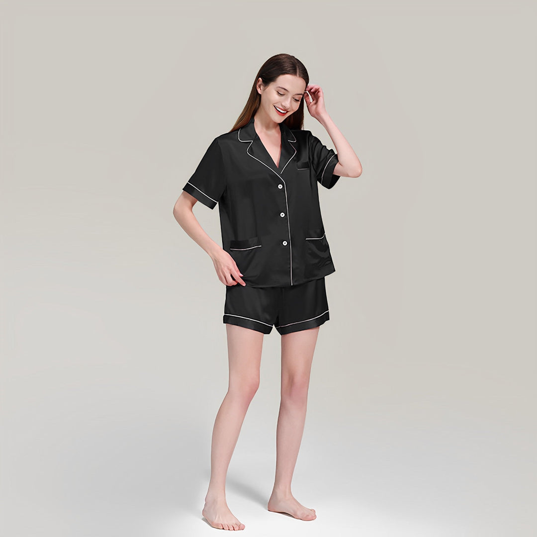 A person stands smiling while wearing the SusanSilk 19 Momme Short Silk Pajamas Set, featuring black silk button-up pajamas with white trim and shorts, against a plain background.