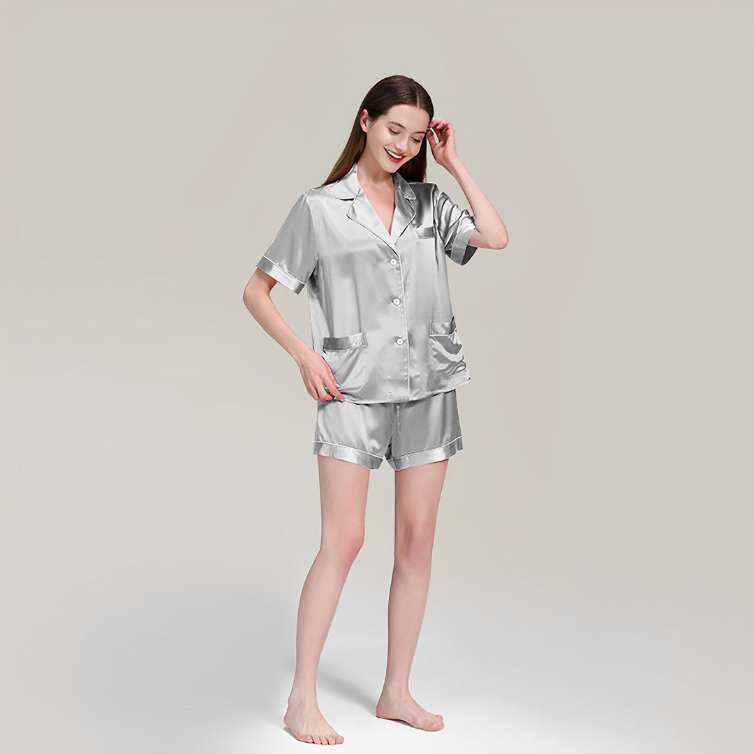 A woman is standing barefoot, smiling, and wearing a 19 Momme Short Silk Pajamas Set by SusanSilk, featuring a short-sleeve button-up top and shorts, against a plain gray background.