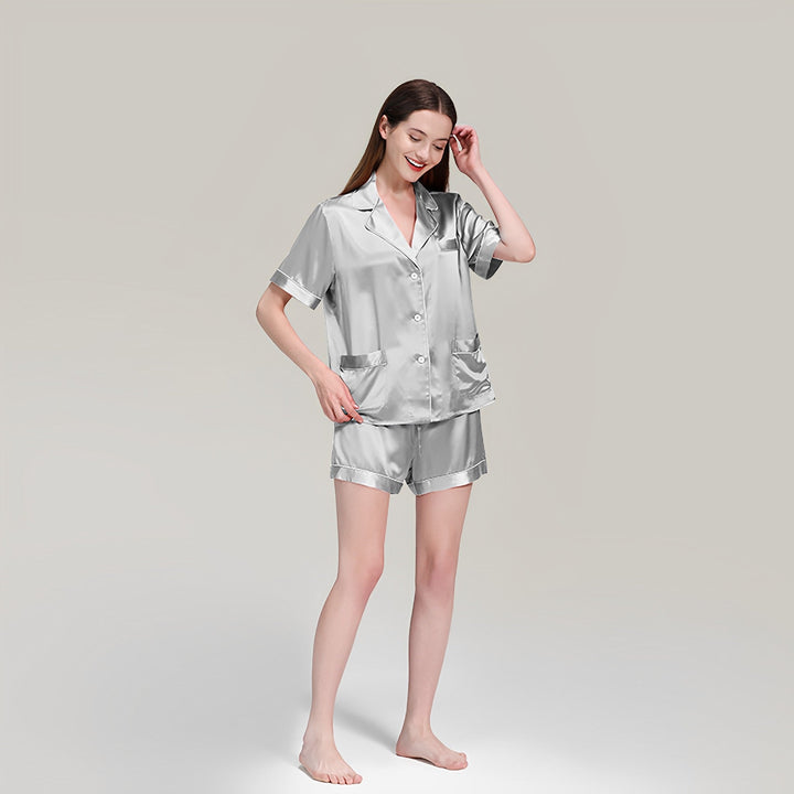 A woman is standing barefoot, smiling, and wearing a 19 Momme Short Silk Pajamas Set by SusanSilk, featuring a short-sleeve button-up top and shorts, against a plain gray background.