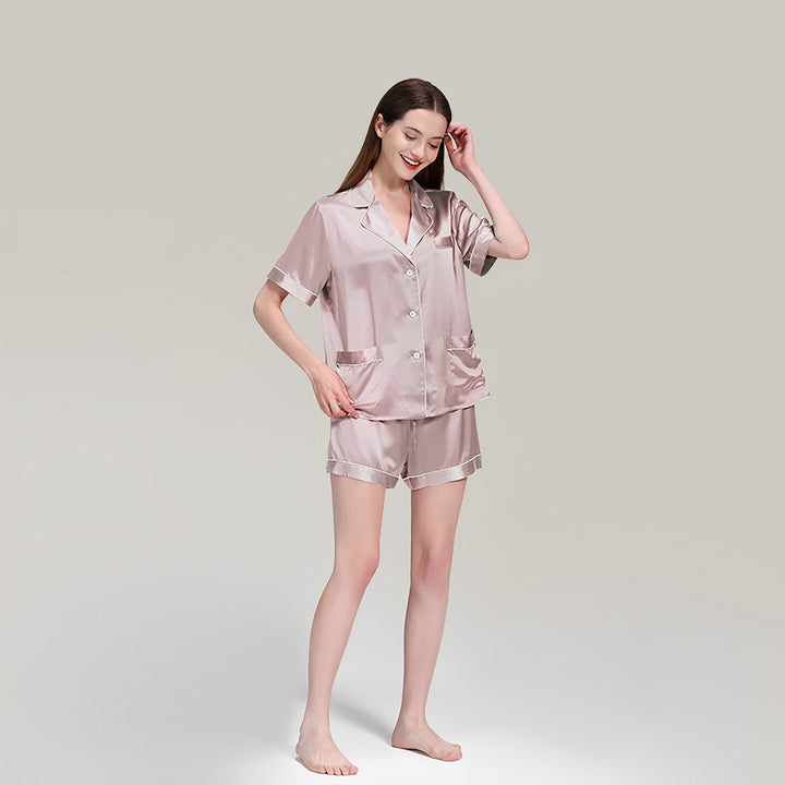 A woman wearing the 19 Momme Short Silk Pajamas Set from SusanSilk, featuring a light pink hue with short sleeves and shorts, stands barefoot on a neutral background, smiling and touching her hair.
