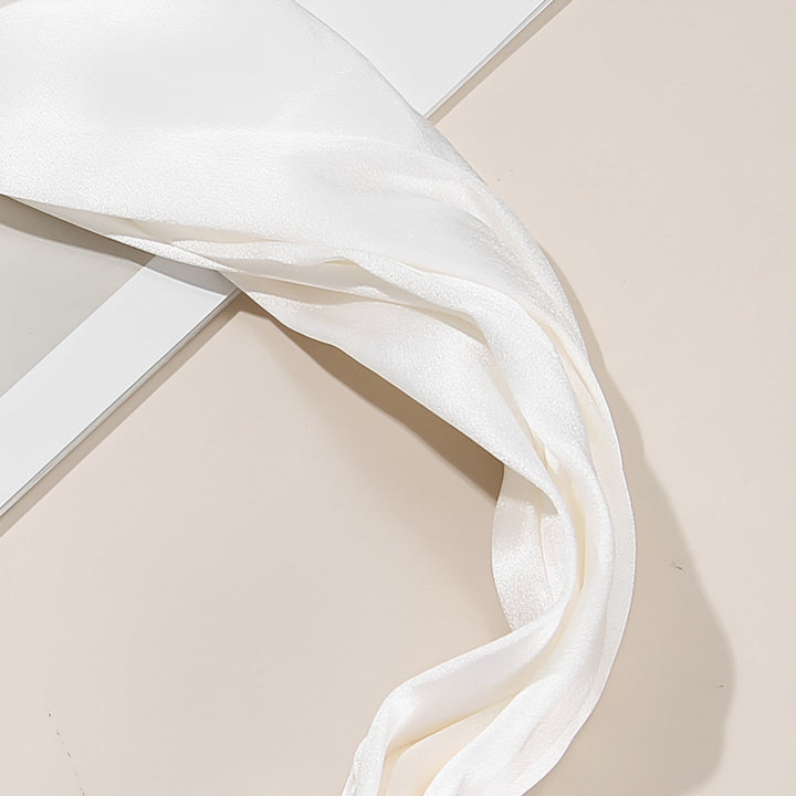 A Pure Color Criss Cross Silk Headband from SusanSilk, reminiscent of delicate satin ribbon, lies partially on a white surface next to a reflective glass surface.