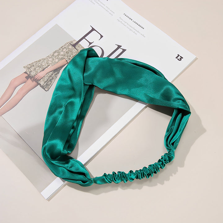 A SusanSilk Pure Color Criss Cross Silk Headband in green is placed on an open magazine showcasing a partially visible fashion image.