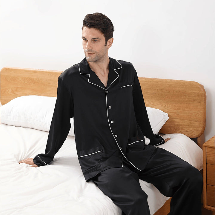 A person in the 19 Momme Lapel Collar Silk Pajamas Set by SusanSilk sits on the edge of a neatly made bed with white bedding and a wooden headboard.
