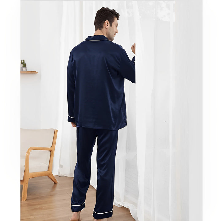 A person wearing SusanSilk's 19 Momme Lapel Collar Silk Pajamas Set in dark blue stands near a sheer curtain in a room with wooden flooring and a wooden chair in the background.