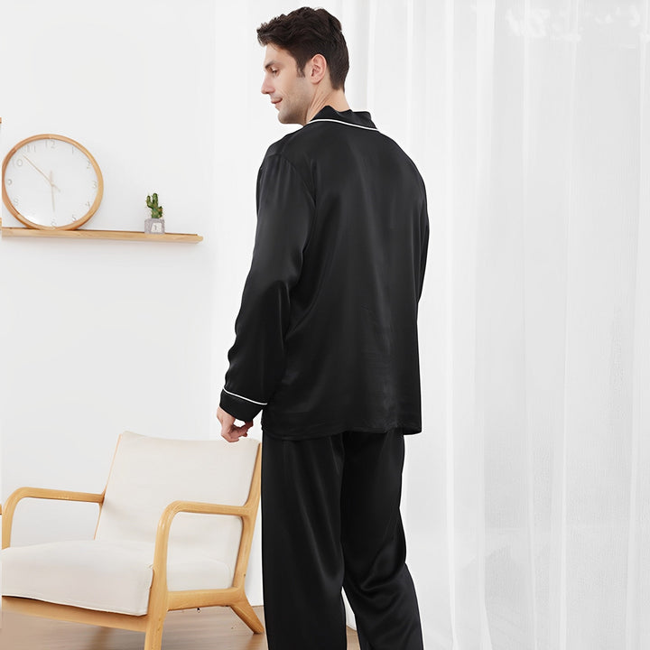 A person dressed in a SusanSilk 19 Momme Lapel Collar Silk Pajamas Set stands near a white armchair and a wooden shelf with a clock and plant.