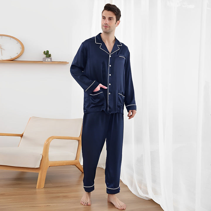 A man stands in a room wearing the 19 Momme Lapel Collar Silk Pajamas Set from SusanSilk. He is near a wooden chair with a wall shelf in the background.