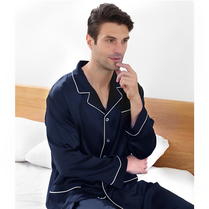 A person wearing the 19 Momme Lapel Collar Silk Pajamas Set from SusanSilk in dark blue with white piping sits on a bed, touching their chin with one hand and crossing their arm, appearing thoughtful.