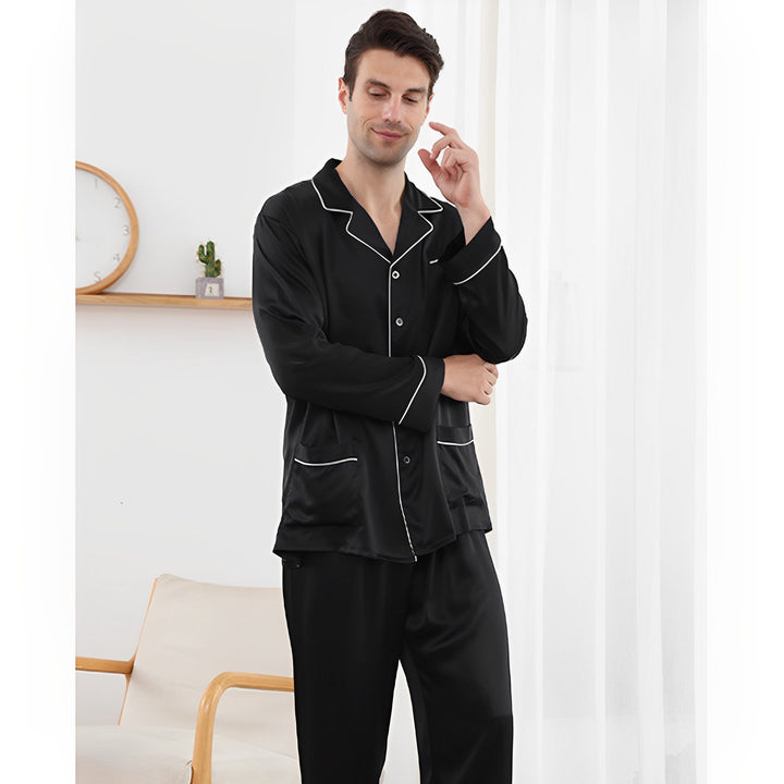 A person in a SusanSilk 19 Momme Lapel Collar Silk Pajamas Set stands next to a chair and a white curtain, touching their face thoughtfully.
