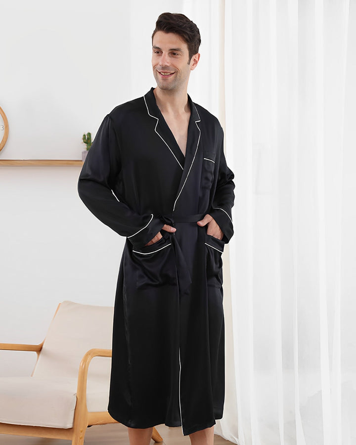 An individual indoors, adorned in a SusanSilk 19 Momme Piping Silk Robe featuring black silk with white trim, stands with hands in pockets and a smile on their face. In the background are a window draped with white curtains and a chair.