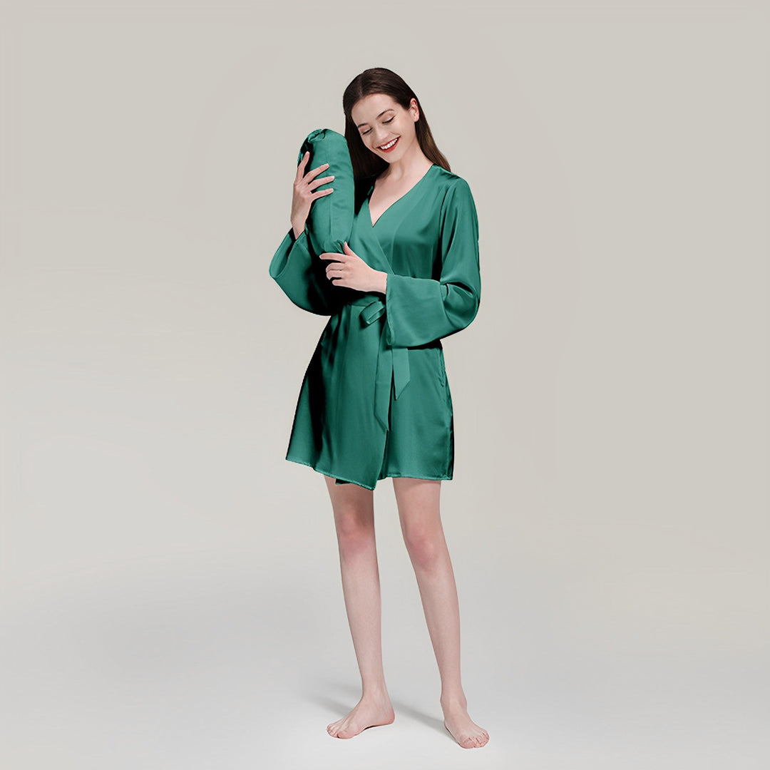A woman in a 19 Momme Mid Length Silk Robe from SusanSilk stands barefoot, smiling with her eyes closed, and holds a SusanSilk pillow close to her face.