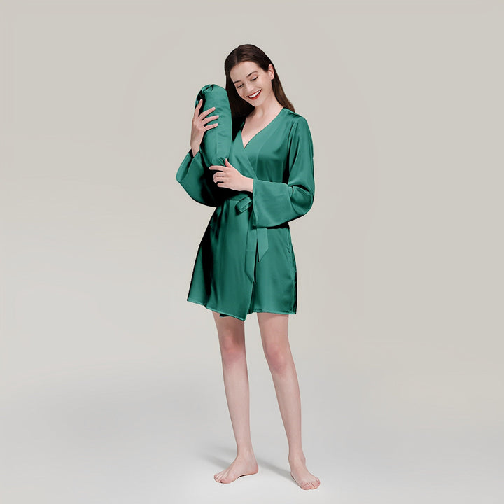 A woman in a 19 Momme Mid Length Silk Robe from SusanSilk stands barefoot, smiling with her eyes closed, and holds a SusanSilk pillow close to her face.