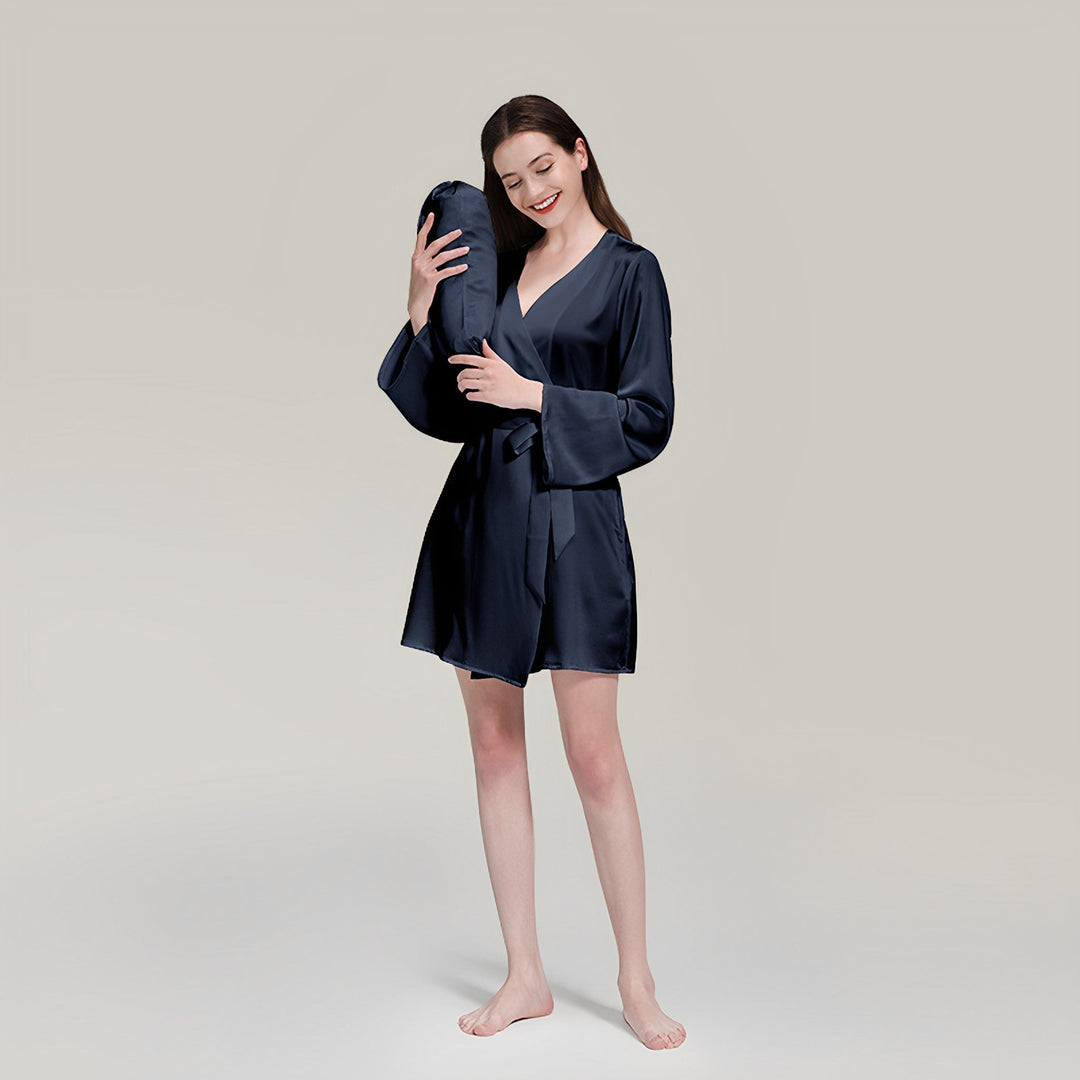 A woman in a navy 19 Momme Mid Length Silk Robe by SusanSilk stands barefoot, holding a matching navy pillow with a content expression. She is set against a plain background.