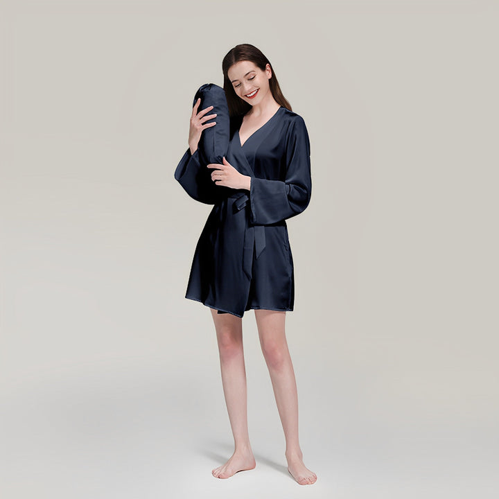 A woman in a navy 19 Momme Mid Length Silk Robe by SusanSilk stands barefoot, holding a matching navy pillow with a content expression. She is set against a plain background.