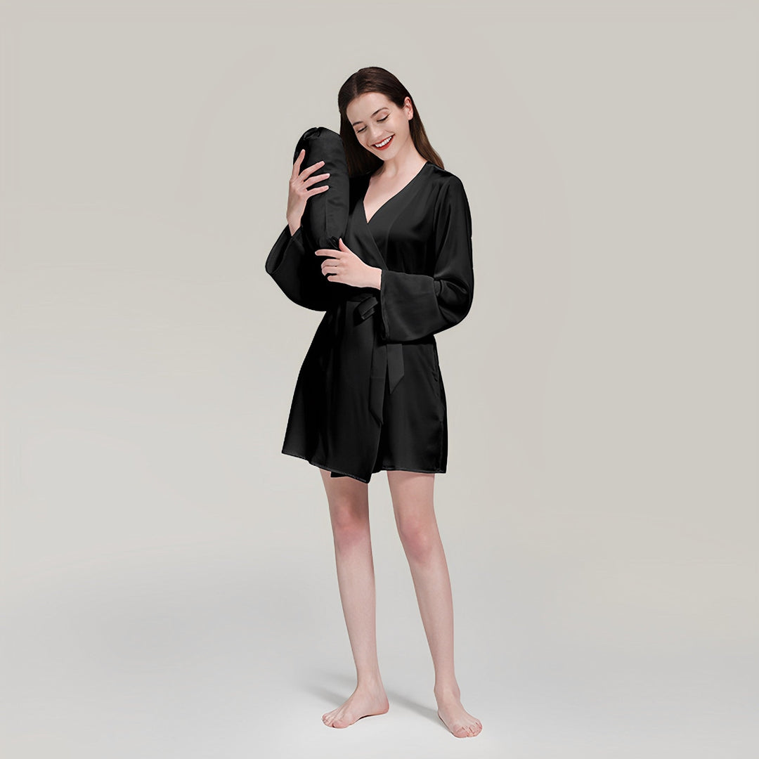 A woman in a 19 Momme Mid Length Silk Robe by SusanSilk stands barefoot, holding a silky black pillow to her head with a relaxed expression on a neutral background.