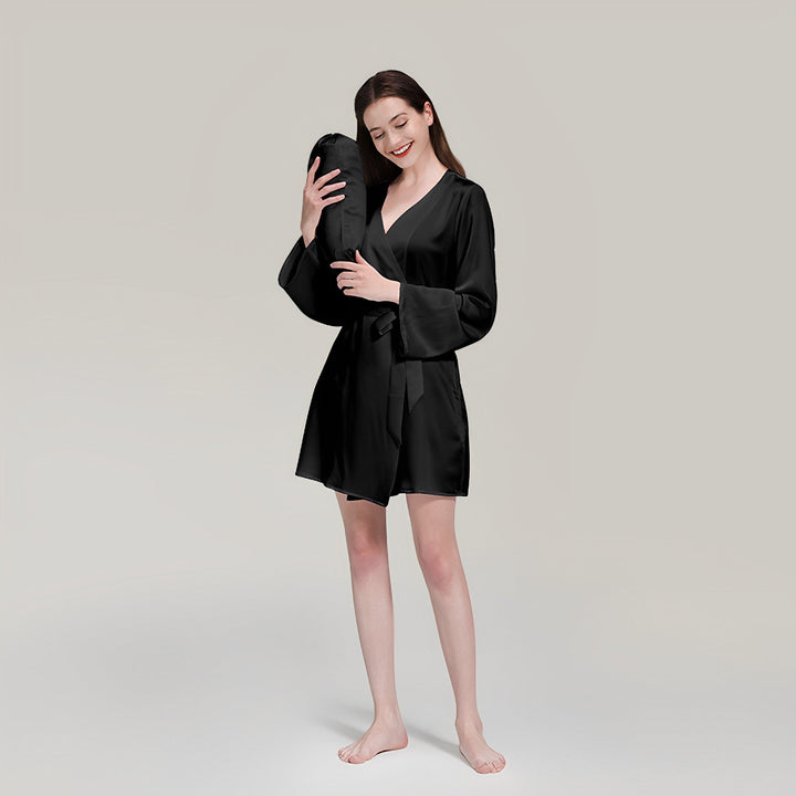 A woman in a 19 Momme Mid Length Silk Robe by SusanSilk stands barefoot, holding a silky black pillow to her head with a relaxed expression on a neutral background.