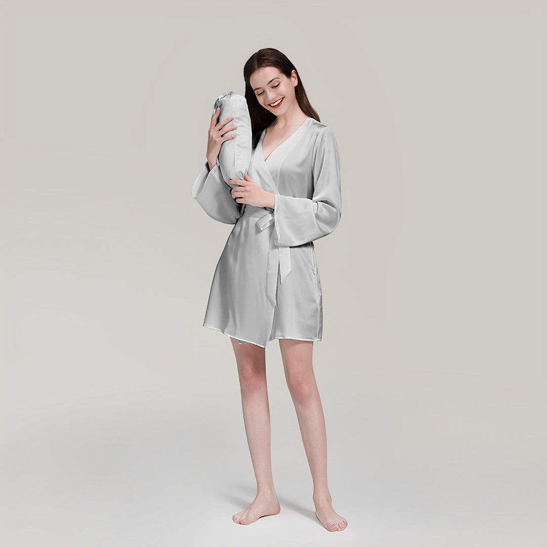 A woman wearing a SusanSilk 19 Momme Mid Length Silk Robe stands barefoot on a white surface, holding a matching silver body pillow, and smiling with her eyes closed.