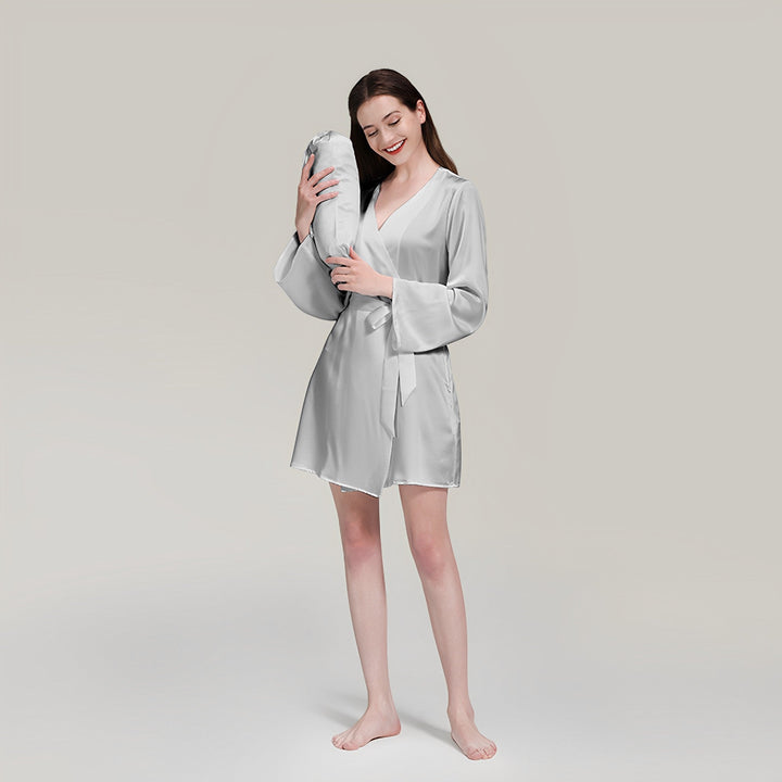 A woman wearing a SusanSilk 19 Momme Mid Length Silk Robe stands barefoot on a white surface, holding a matching silver body pillow, and smiling with her eyes closed.