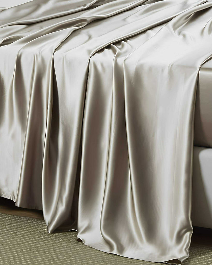 A bed is draped with the luxurious 19 Momme Moisturizing Silk Bedding Set from SusanSilk, featuring smooth, shiny, silver-colored sheets that cascade down the sides in soft, elegant folds.