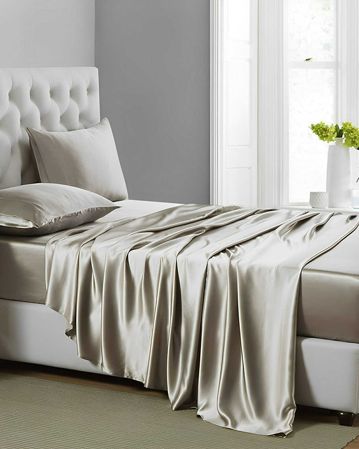 A neatly made bed adorned with the luxurious SusanSilk 19 Momme Moisturizing Silk Bedding Set - 4Pcs in silver satin is set in a modern bedroom featuring a tufted gray headboard. Sunlight streams through the window, illuminating a small white side table topped with green flowers.