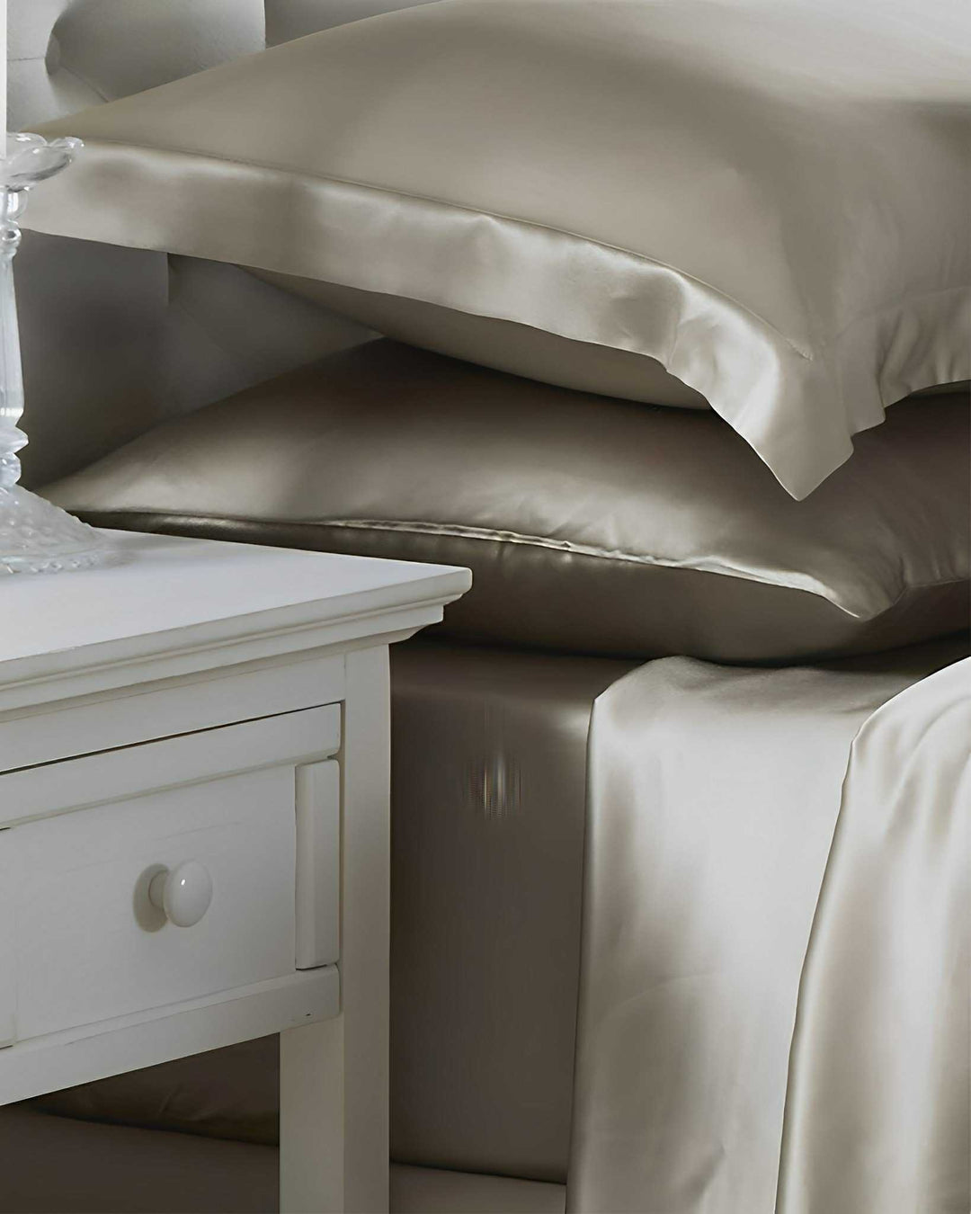 A neatly made bed adorned with the 19 Momme Moisturizing Silk Bedding Set - 4Pcs by SusanSilk in beige, complete with silk satin sheets and pillows, sits next to a white bedside table featuring a glass lamp.