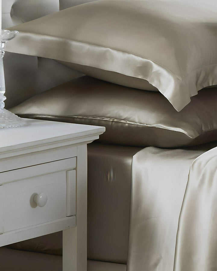 A neatly made bed adorned with the 19 Momme Moisturizing Silk Bedding Set - 4Pcs by SusanSilk in beige, complete with silk satin sheets and pillows, sits next to a white bedside table featuring a glass lamp.