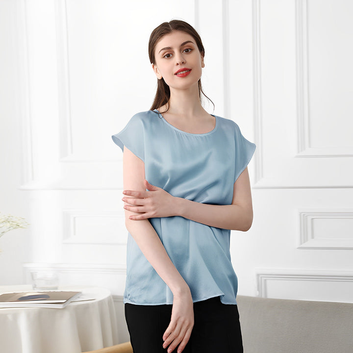 A woman stands indoors, wearing the SusanSilk Short Sleeves Round Neck Silk Tee in light blue and black pants, with her arms loosely crossed over her torso. She is facing the camera and smiling slightly.