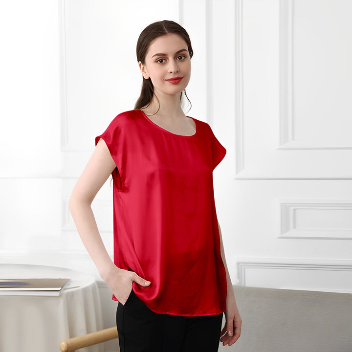 A woman wearing a SusanSilk Short Sleeves Round Neck Silk Tee in red and black pants stands indoors, posed with one hand in her pocket. She is positioned against a white paneled wall with a table and chair visible in the background.