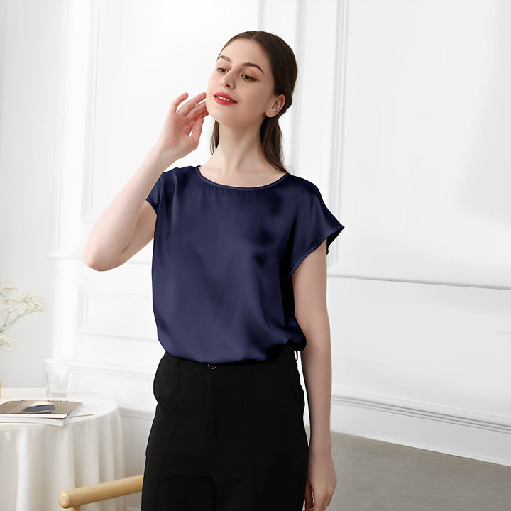 A woman wearing a navy blue Short Sleeves Round Neck Silk Tee by SusanSilk and black pants stands indoors, touching her face with one hand. A table with a book, glass, and plant is in the background.