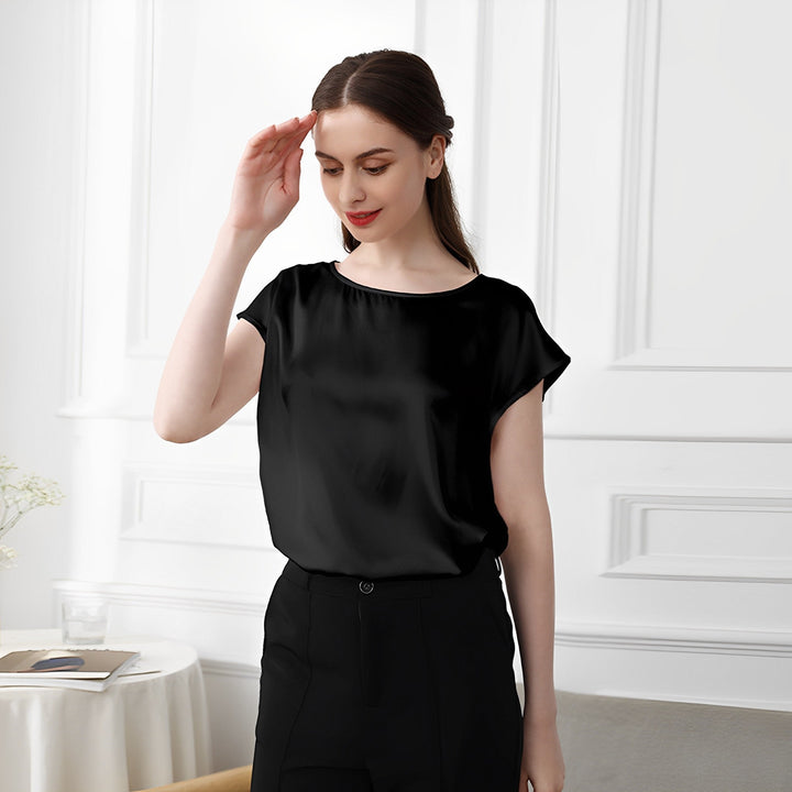 A woman in a Short Sleeves Round Neck Silk Tee by SusanSilk and black pants stands in a room with white walls and a small round table. She is looking down and touching her hair with one hand.