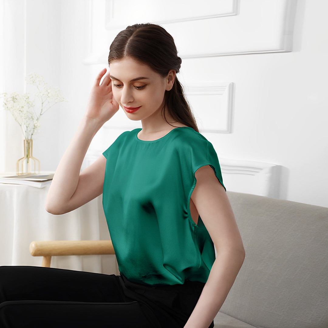 A woman in a SusanSilk Short Sleeves Round Neck Silk Tee and black pants sits on a sofa, touching her hair with one hand, in a well-lit room with white walls and minimal decor.
