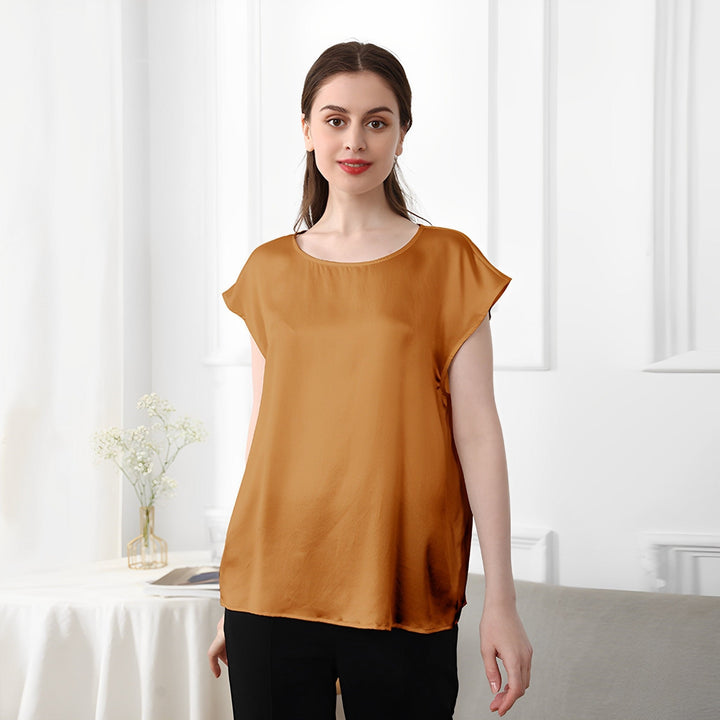 A woman stands in a room wearing a mustard yellow SusanSilk Short Sleeves Round Neck Silk Tee and black pants, with a small table and a vase of flowers in the background.