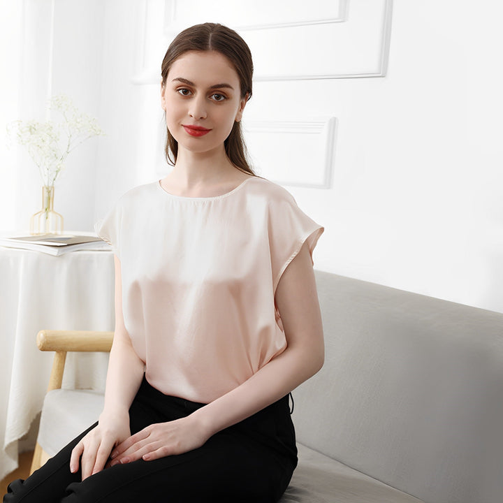 A woman wearing a light-colored SusanSilk Short Sleeves Round Neck Silk Tee and black pants is seated on a gray couch in a modern, minimalist room with white walls and a small table adorned with a vase of flowers.