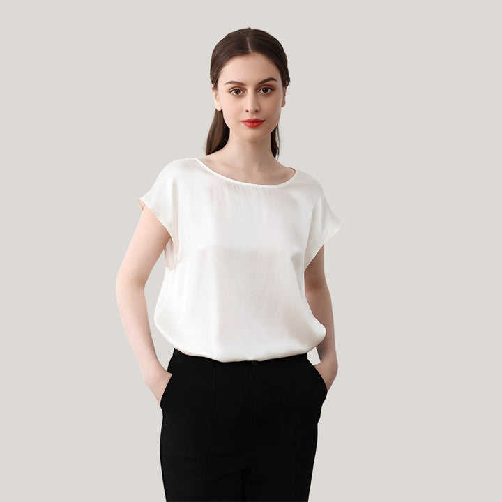 A woman in a Short Sleeves Round Neck Silk Tee by SusanSilk and black pants stands against a plain, light-colored background. Her hands are in her pockets, and she has a neutral expression.
