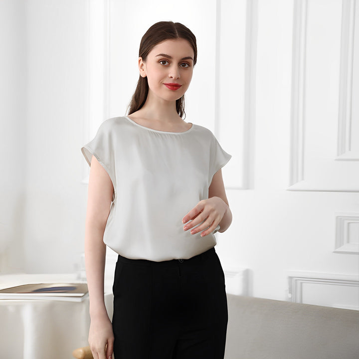A woman with long brown hair, wearing a white SusanSilk Short Sleeves Round Neck Silk Tee and black pants, stands indoors, smiling softly. The background features white walls and a partial view of wooden furniture.