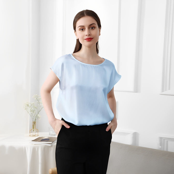 A woman stands in a well-lit room, wearing a SusanSilk Short Sleeves Round Neck Silk Tee in light blue and black pants. She has shoulder-length brown hair and a neutral expression. A table with a vase of flowers is in the background.