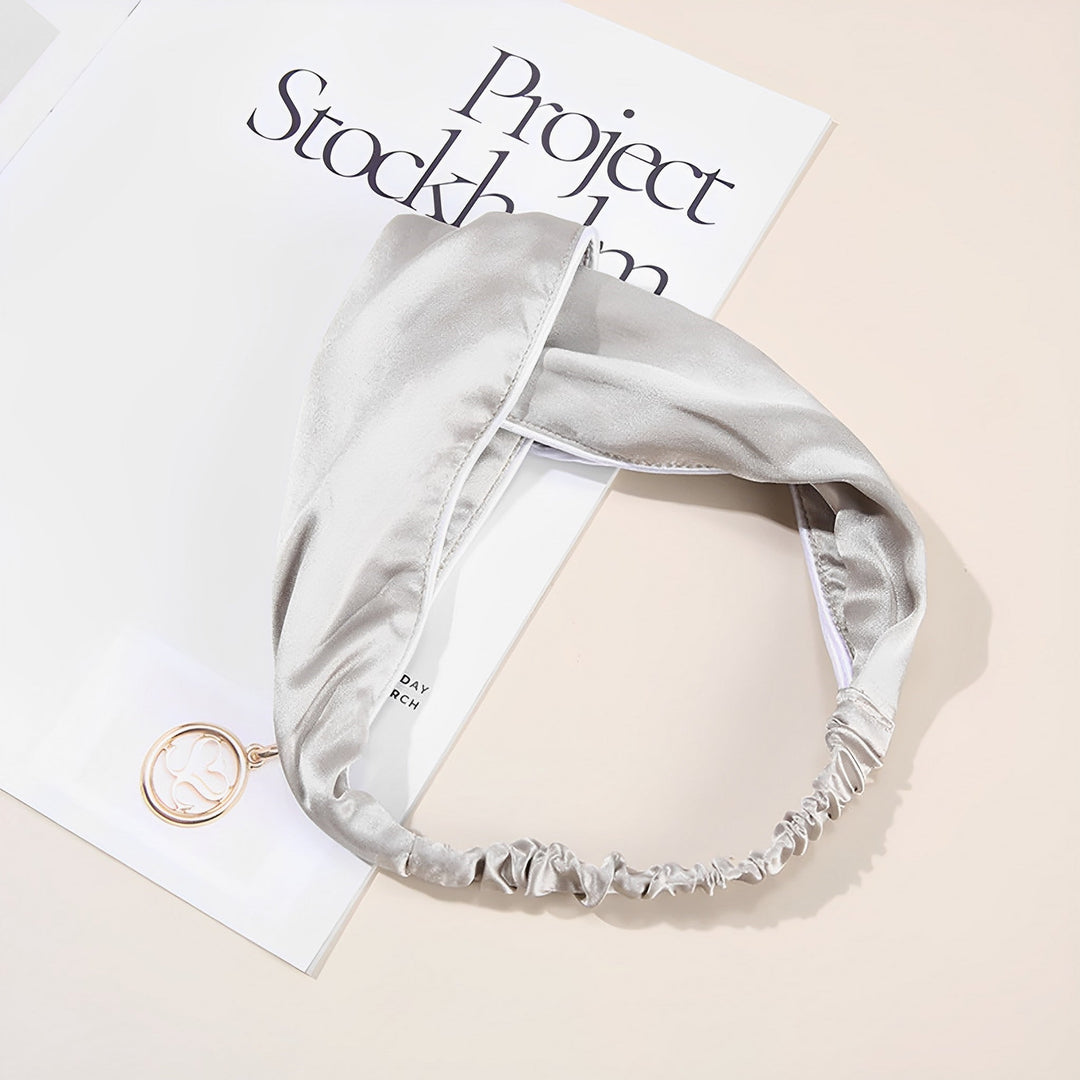 A SusanSilk Piping Criss Cross Silk Headband is placed on an open magazine titled "Project Stockholm." Alongside, a round pendant with a tree design lies on the magazine, adding elegance to the serene scene.